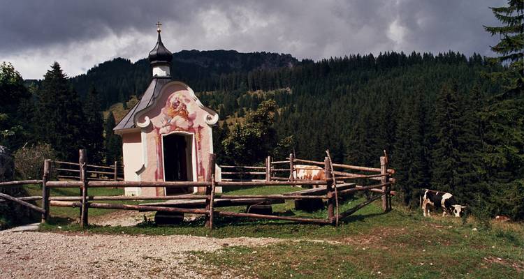 Pequeña capilla rodeada por una cerca de madera, con vacas pastando cerca y un bosque denso al fondo.