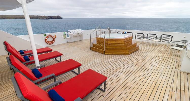 Zonnedek met rode ligstoelen en een houten hottub met uitzicht op open oceaan onder lichte bewolking.