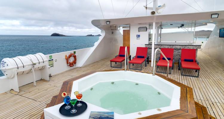 Open deck of a yacht featuring a small jacuzzi, red lounge chairs, and ocean backdrop.