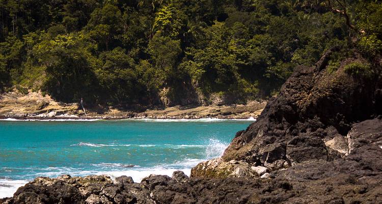 Las olas turquesas se estrellan contra rocas volcánicas oscuras con una densa selva tropical al fondo.