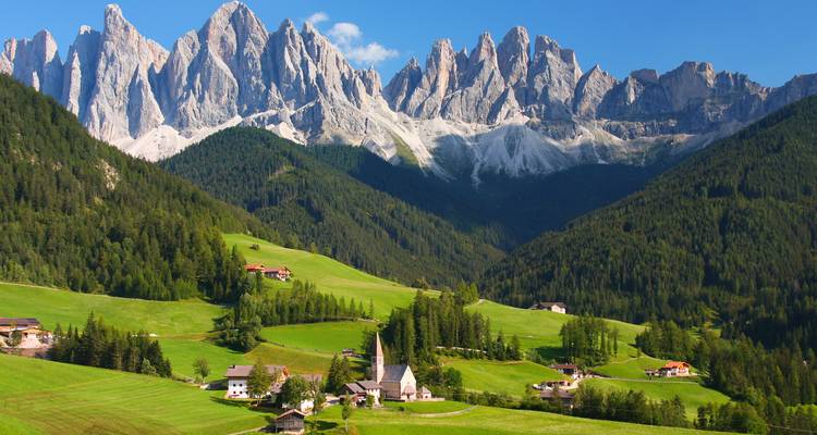Idyllisches Alpental mit grünen Wiesen und Kirche unter den gezackten Dolomitengipfeln unter blauem Himmel.