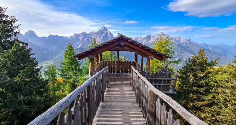 Eine hölzerne Aussichtsbrücke führt zur imposanten Dolomiten-Gebirgskette unter einem lebendigen blauen Himmel.