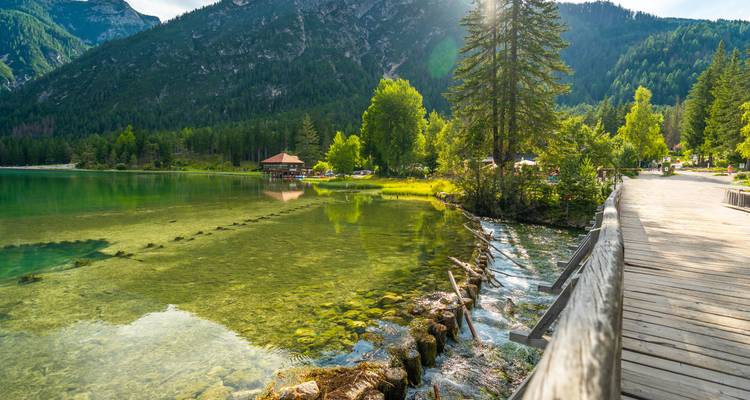 Kristallklarer Bergsee, umgeben von Kiefern und Bergen, mit einem hölzernen Gehweg, der entlang des Ufers führt.