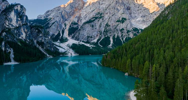 Luftaufnahme des türkisfarbenen Lago di Braies, der raue Felsklippen und dichten Kiefernwald widerspiegelt.