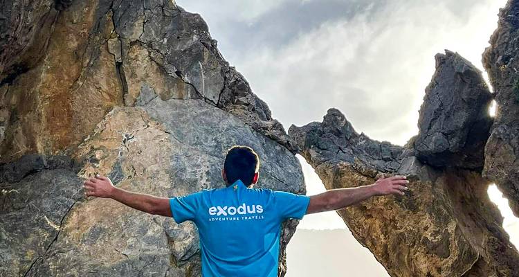 Viajero con los brazos extendidos celebrando bajo un arco de roca irregular contra un cielo dramático
