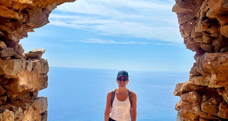 Mujer sonriente enmarcada por una ventana de piedra arruinada muy por encima de un profundo mar Mediterráneo azul