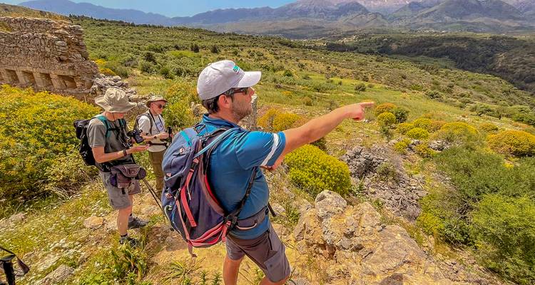 Un guía señalando a través de un valle soleado mientras dos senderistas observan, con mochilas y bastones listos