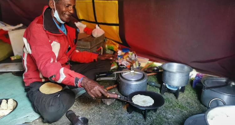 El cocinero del campamento prepara chapati sobre una llama dentro de una tienda de expedición colorida llena de suministros.