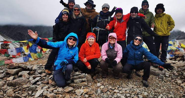 Un grand groupe de trekkeurs vêtus de vestes colorées pose parmi les drapeaux de prière sur un col de montagne rocheux sous un ciel nuageux.