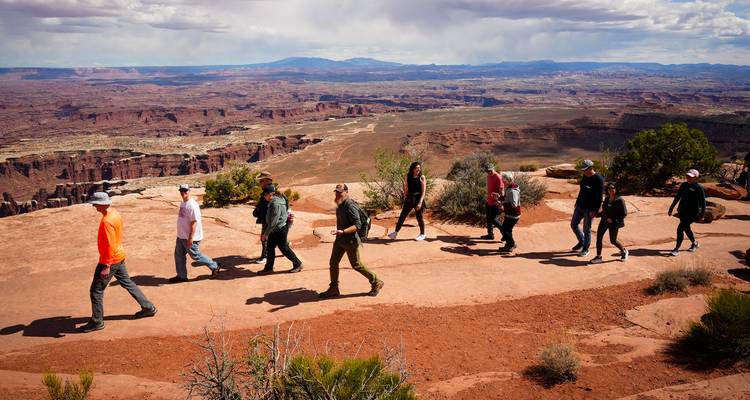 Line of hikers walks a red rock rim overlooking vast canyon vistas and distant mesas.