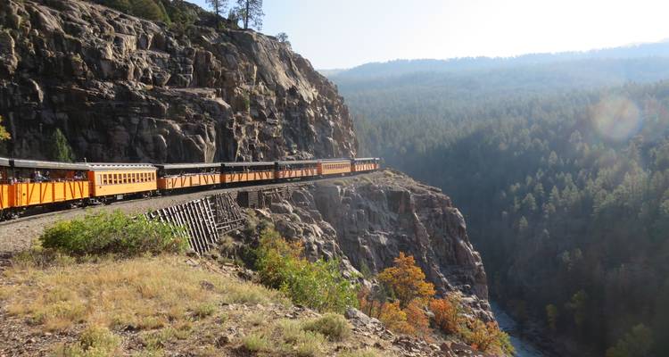 Train historique à voie étroite jaune et noir traverse un pont en treillis spectaculaire au bord d'une falaise au-dessus d'un canyon boisé.