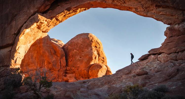 Randonneur solitaire en silhouette sous une imposante arche naturelle de grès rouge à l'aube.