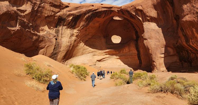 Des voyageurs marchent à travers un canyon sablonneux vers une immense arche avec un trou circulaire haut dans la paroi.
