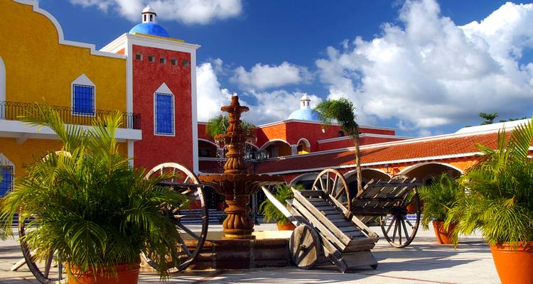 Patio colonial colorido con fuente, ruedas de carreta y edificios amarillo brillante y rojo bajo un cielo azul.