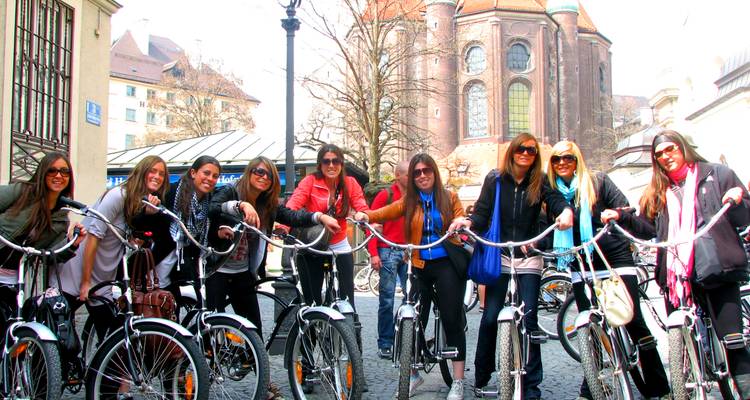 Un groupe joyeux de jeunes femmes sur des vélos urbains brillants posant dans une cour historique de Munich avec une église en arrière-plan.
