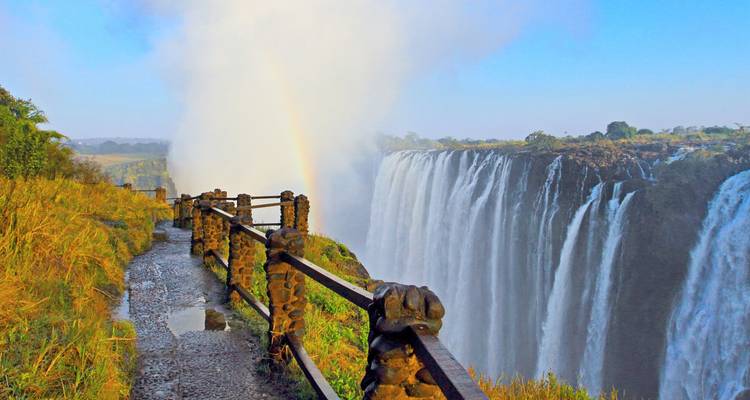 Spectaculair uitzicht op Victoria Falls met hoge mistwalmen en een levendige regenboog naast een stenen voetpad langs de rand van de klif.
