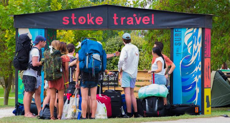 Voyageurs avec des sacs à dos près d'un stand d'information 'Stoke Travel'.
