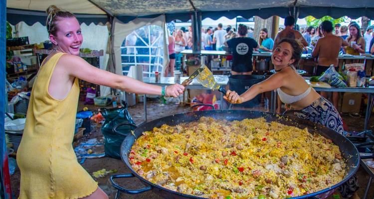 Two women holding large utensils over a giant pan of paella at a festival