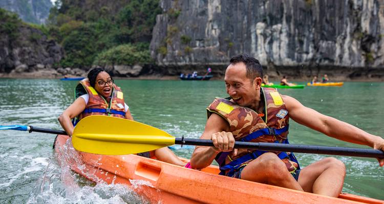 Una pareja emocionada rema en un kayak naranja a través de aguas esmeralda rodeadas por imponentes acantilados de piedra caliza de la Bahía de Halong.