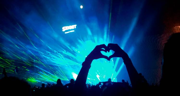 Concertgoers raise a heart-shaped silhouette against laser lights and the glowing SZIGET Festival stage at night.