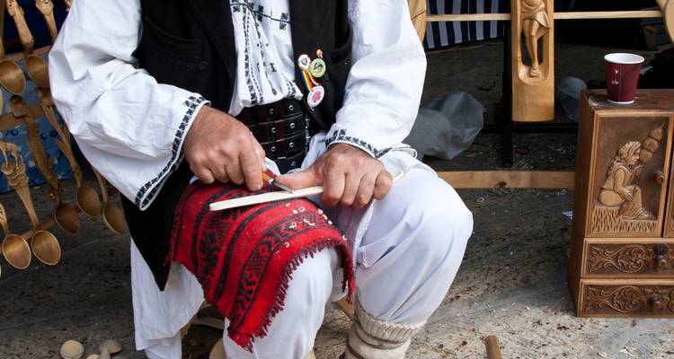 Artisan in traditional attire carving a wooden spoon while seated at a stall displaying crafts