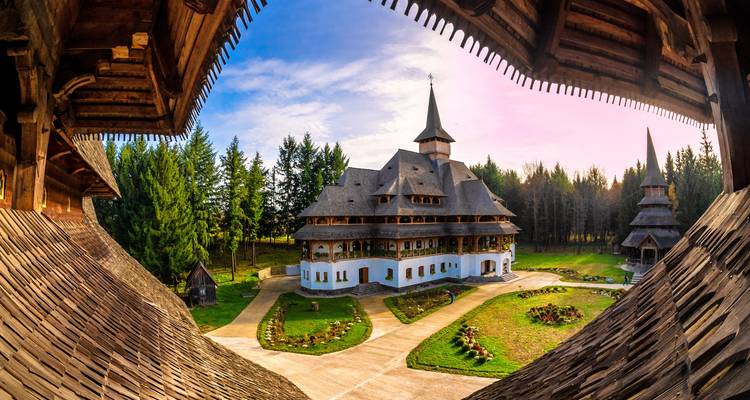 Panoramic framed view of a grand wooden monastery complex amid dense pine forest at sunset