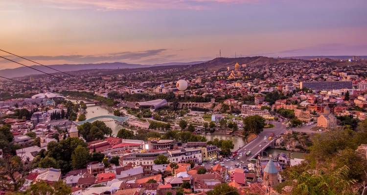 Vista panorámica del atardecer sobre la ciudad de Tiflis con línea de teleférico, río Kura y monumentos iluminados.