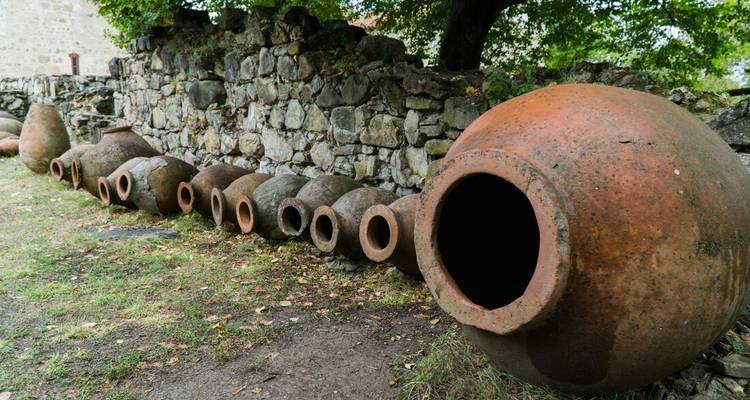 Una hilera de grandes vasijas tradicionales de arcilla qvevri para vino descansando junto a una vieja pared de piedra bajo un árbol