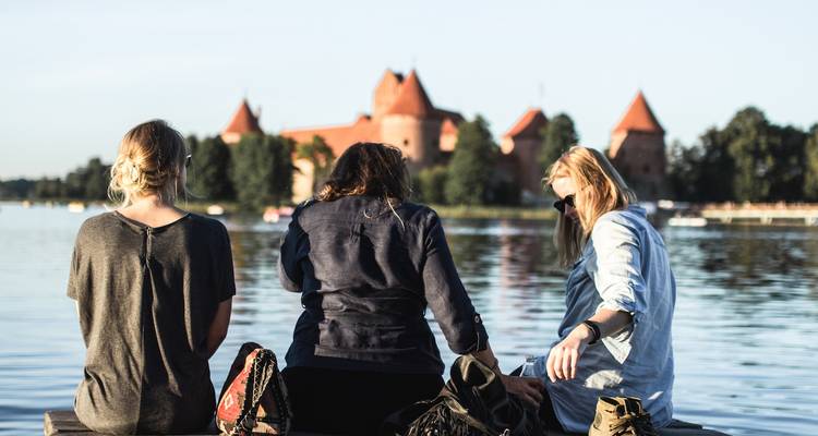 Tres personas sentadas junto al agua con el castillo de la isla de Trakai al fondo.