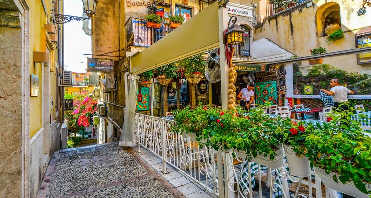 Ruelle étroite et colorée bordée de fleurs et de cafés, avec des touristes dînant en plein air.