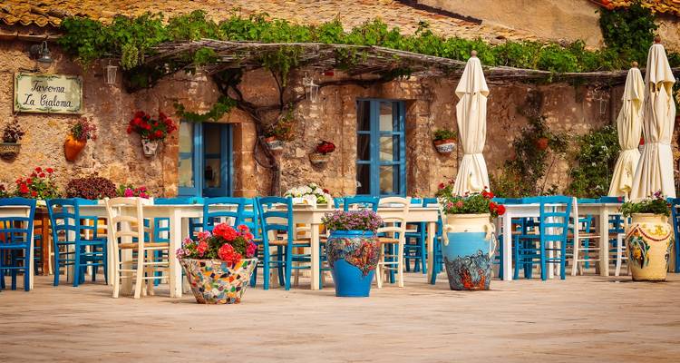 Terrasse de taverne méditerranéenne colorée avec des chaises bleues, des fleurs et des murs en pierre rustique.