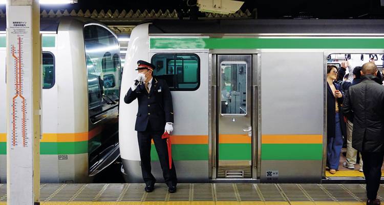 Conductor de tren en uniforme hace señales junto a tren de cercanías con rayas verdes mientras los pasajeros abordan.