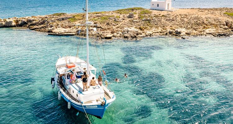 A catamaran anchors in turquoise waters as travellers swim and sunbathe near a small Greek chapel island.