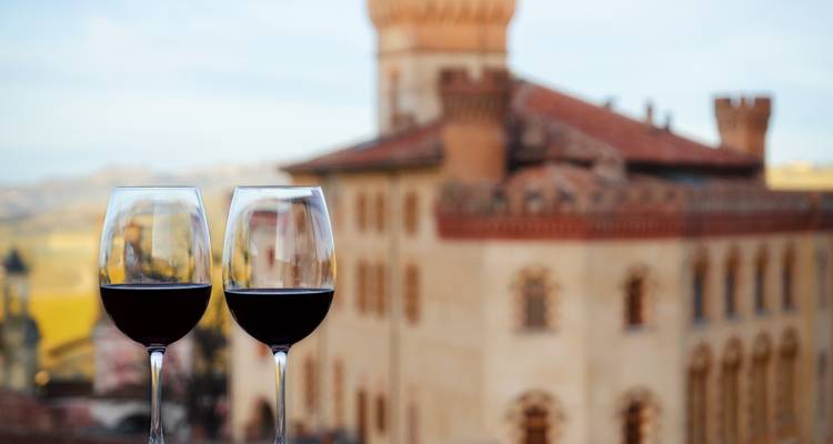 Dos copas de vino tinto chocando con un castillo desenfocado y las colinas ondulantes de Piamonte de fondo