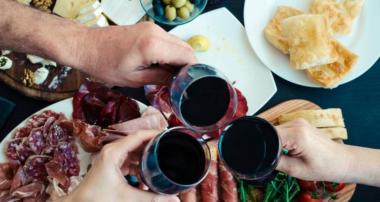 Drei Haende stoßen Glaeser mit Rotwein ueber einem Tisch an, der mit italienischen Wurstwaren, Kaesen, Oliven und Brot gefuellt ist