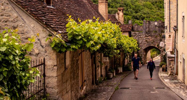 Un callejón de adoquines bordeado de casitas de piedra cubiertas de hiedra y un arco antiguo recibe a dos paseantes disfrutando de un tranquilo paseo por el pueblo.