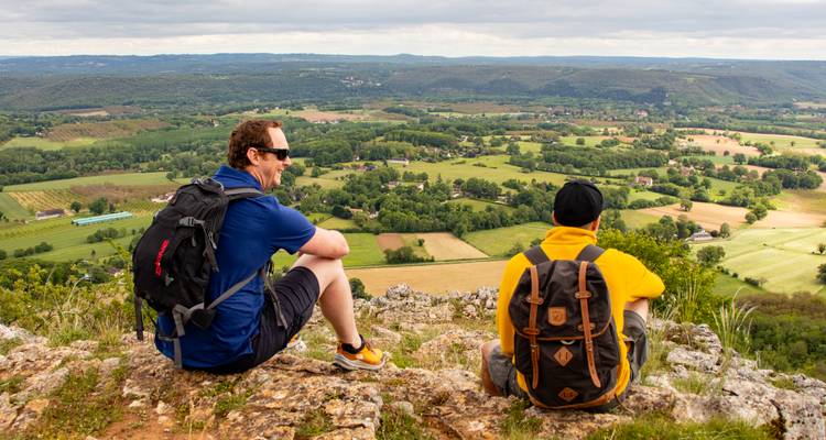 Dos excursionistas se sientan en el borde de un acantilado rocoso mirando hacia un mosaico de verdes valles, granjas y colinas distantes bajo un cielo nublado.