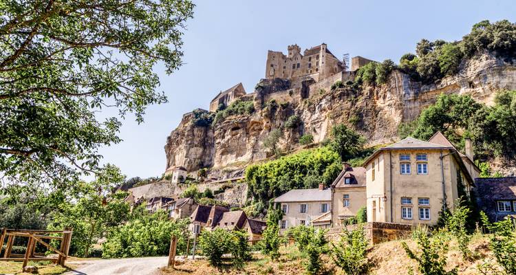 Las casas de piedra se agrupan bajo la dramática fortaleza en lo alto del acantilado de Beynac que se eleva sobre la exuberante vegetación en un día soleado.