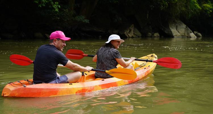 Zwei Reisende paddeln in einem orangen Kajak auf einem ruhigen grünen Fluss, umgeben von bewaldeten Klippen.