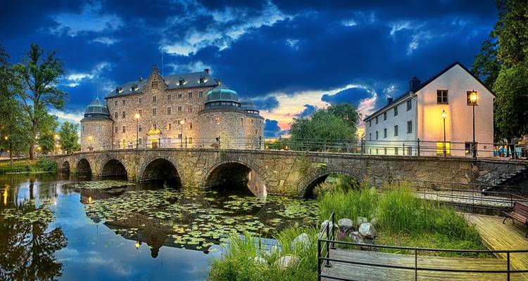 Un castillo histórico con múltiples torres y un puente de piedra sobre un río lleno de nenúfares bajo un cielo dramático.
