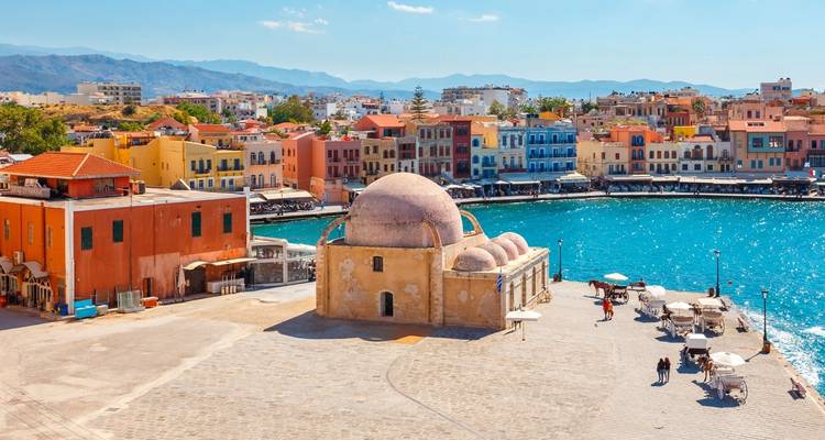 Coloridos edificios portuarios venecianos y la Mezquita de los Jenízaros con cúpula en Chania enmarcando aguas turquesas.