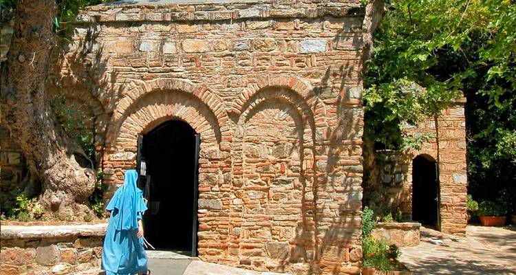 Pequeño santuario de ladrillo rodeado de vegetación con un visitante vestido con túnica entrando por la puerta arqueada.