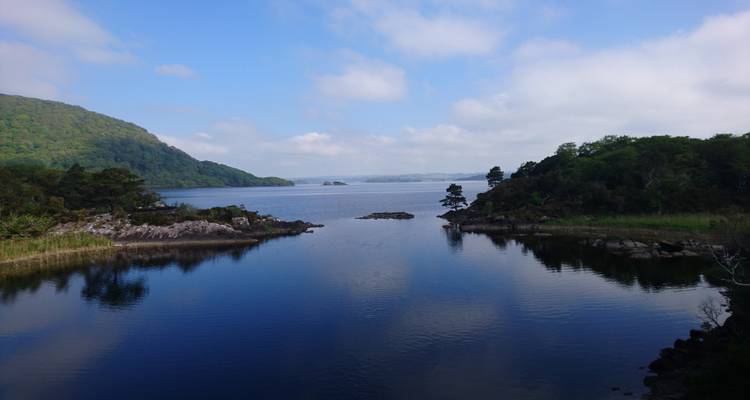 Serene freshwater lake bordered by wooded hillsides reflects a blue sky with scattered clouds