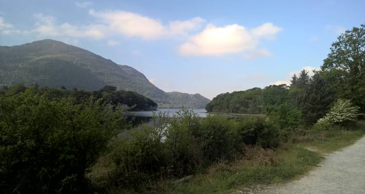 Sentier de gravier au bord d'un lac irlandais paisible avec des collines boisées et des montagnes au loin.