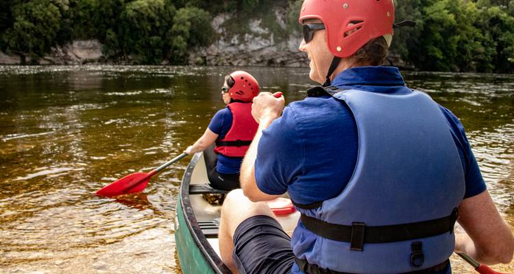 Deux personnes pagayant en canoe sur une rivière aux eaux calmes, portant des casques et des gilets de sauvetage.