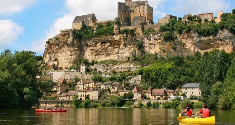 Des personnes font du kayak sur une rivière avec un château à flanc de falaise en arrière-plan.