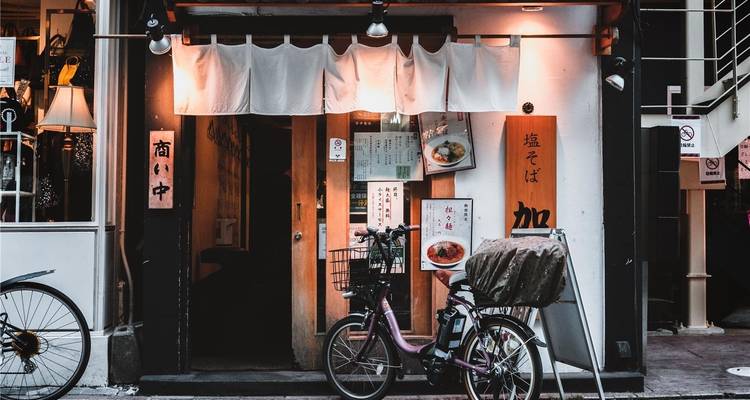 Un acogedor restaurante japonés con cortinas noren blancas y una bicicleta estacionada invita a los transeúntes por una calle estrecha.