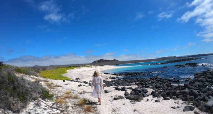 Persoon die langs een zandig strand loopt met turquoise water en rotsachtige kustlijn.