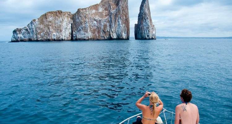 Two people on a boat approaching large sea cliffs in a calm ocean.
