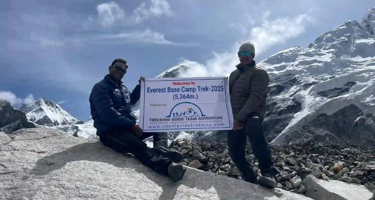 Deux randonneurs tiennent une bannière du Camp de Base de l'Everest au milieu de pics enneigés et d'un terrain rocheux.
